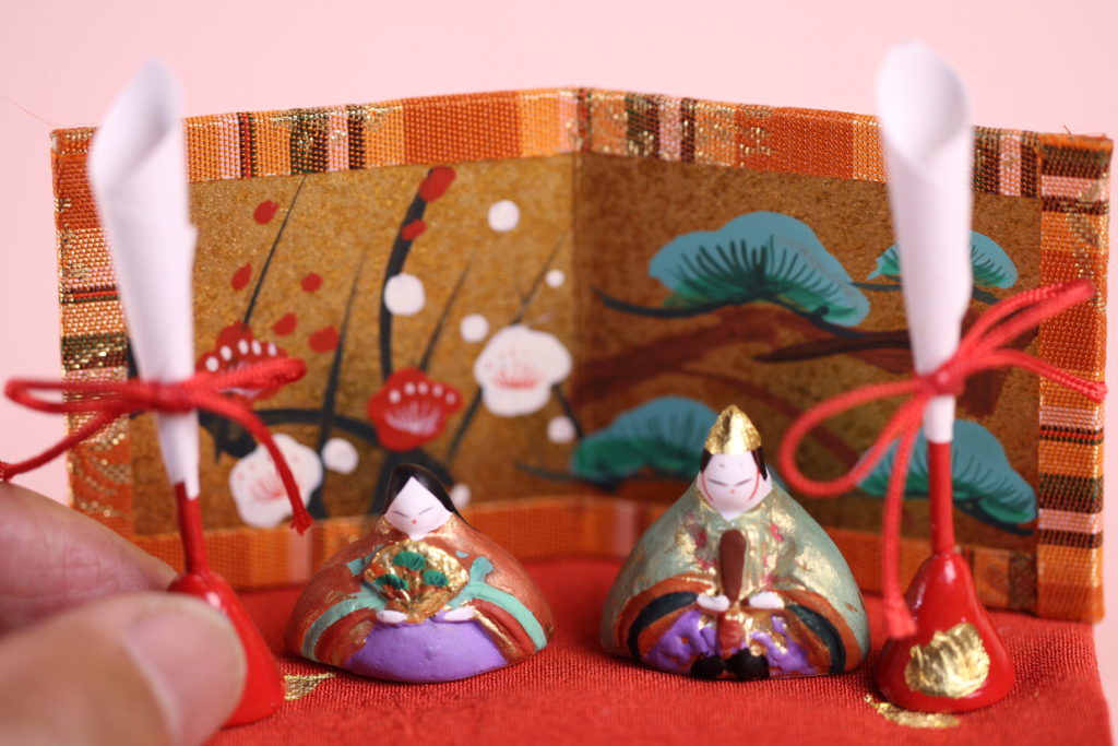 内裏雛の拡大写真です。手にしている油灯も土と江戸組み紐を使って丁寧に手作りしています。もちろん屏風も１枚１枚手描き彩色して、京都から取り寄せた緞子を周りに巻いています。